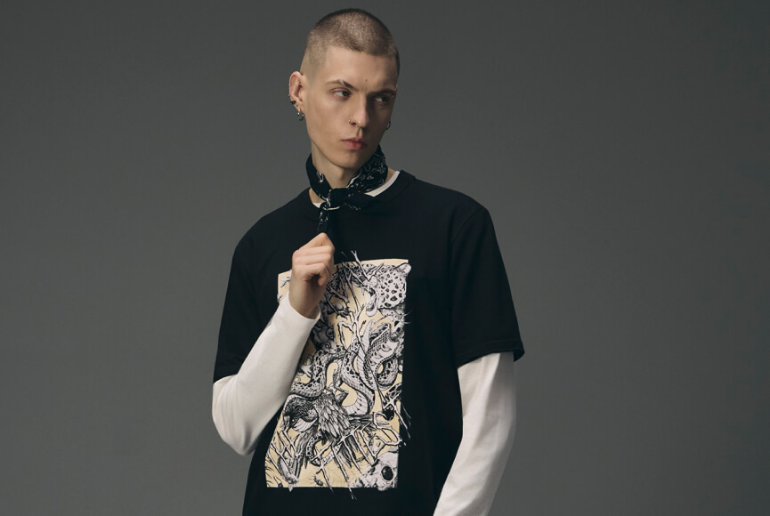 Mężczyzna z krótkimi włosami i kolczykami, ubrany w czarny t-shirt z wyrazistym nadrukiem węża i czaszki założony na biały longsleeve, z czarną bandanką na szyi na szarym tle.