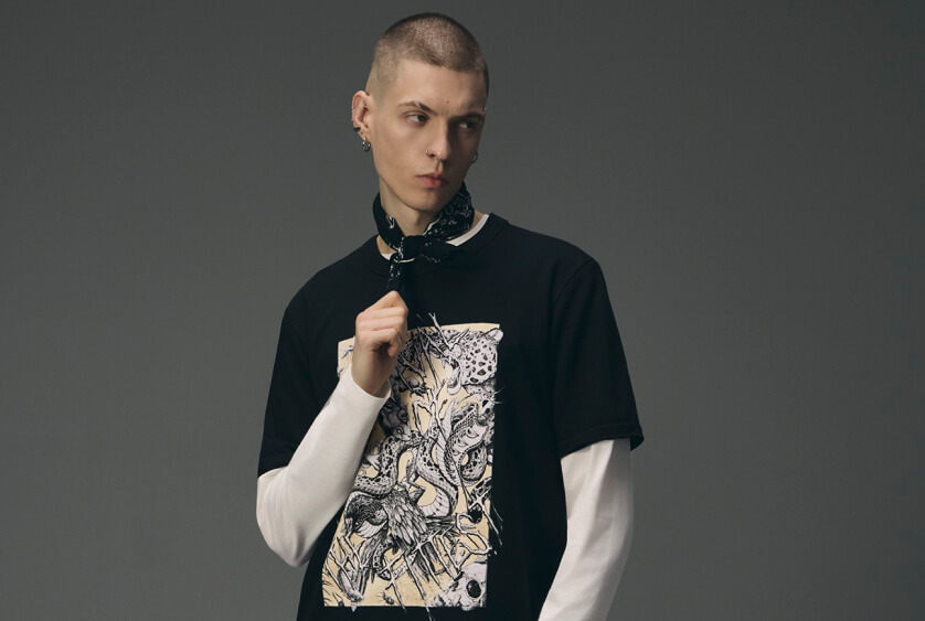 Mężczyzna z krótkimi włosami i kolczykami, ubrany w czarny t-shirt z wyrazistym nadrukiem węża i czaszki założony na biały longsleeve, z czarną bandanką na szyi na szarym tle.