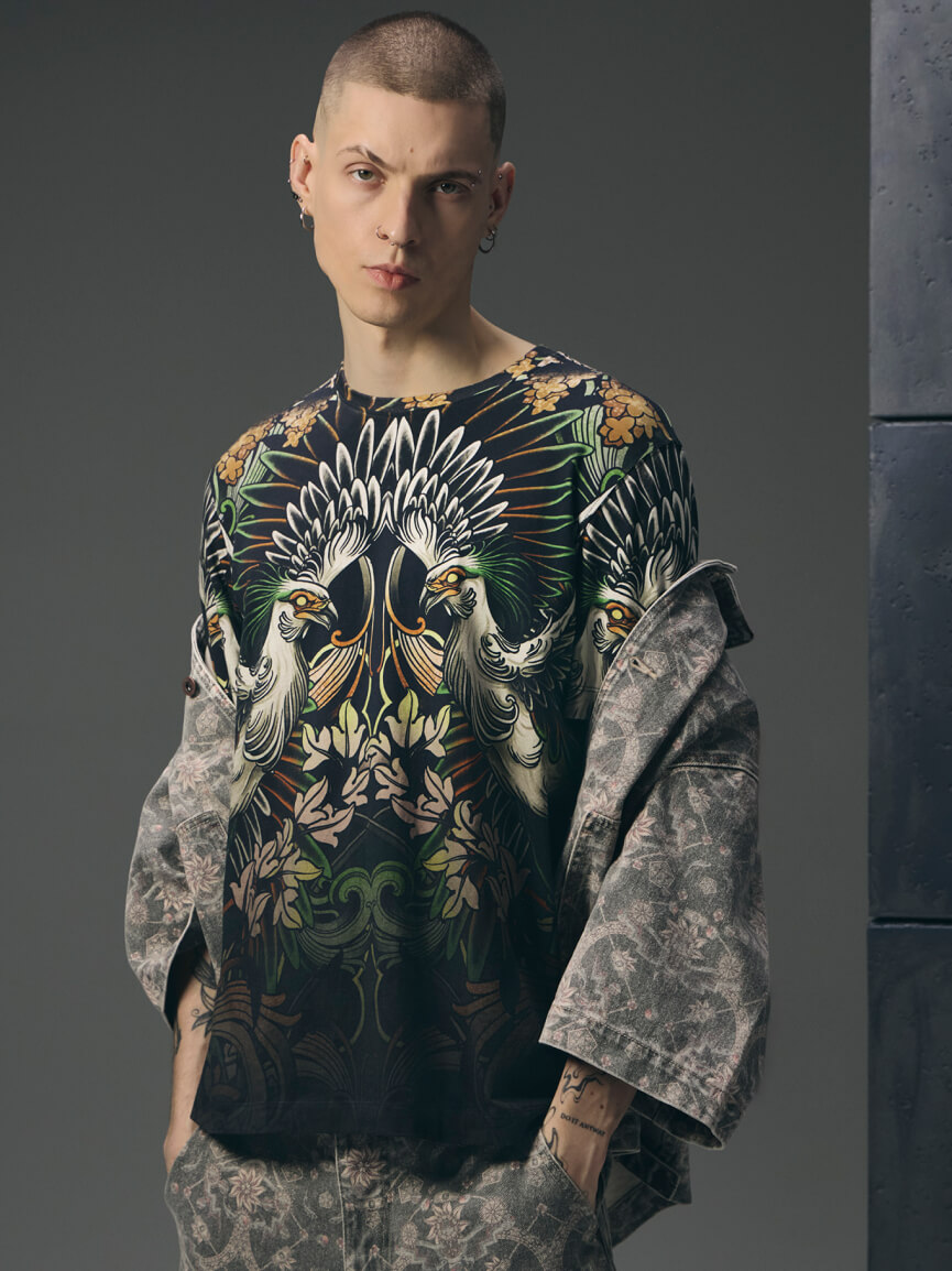 Mężczyzna z krótkimi włosami i kolczykami, ubrany w czarny t-shirt z wyrazistym nadrukiem węża i czaszki założony na biały longsleeve, z czarną bandanką na szyi na szarym tle.