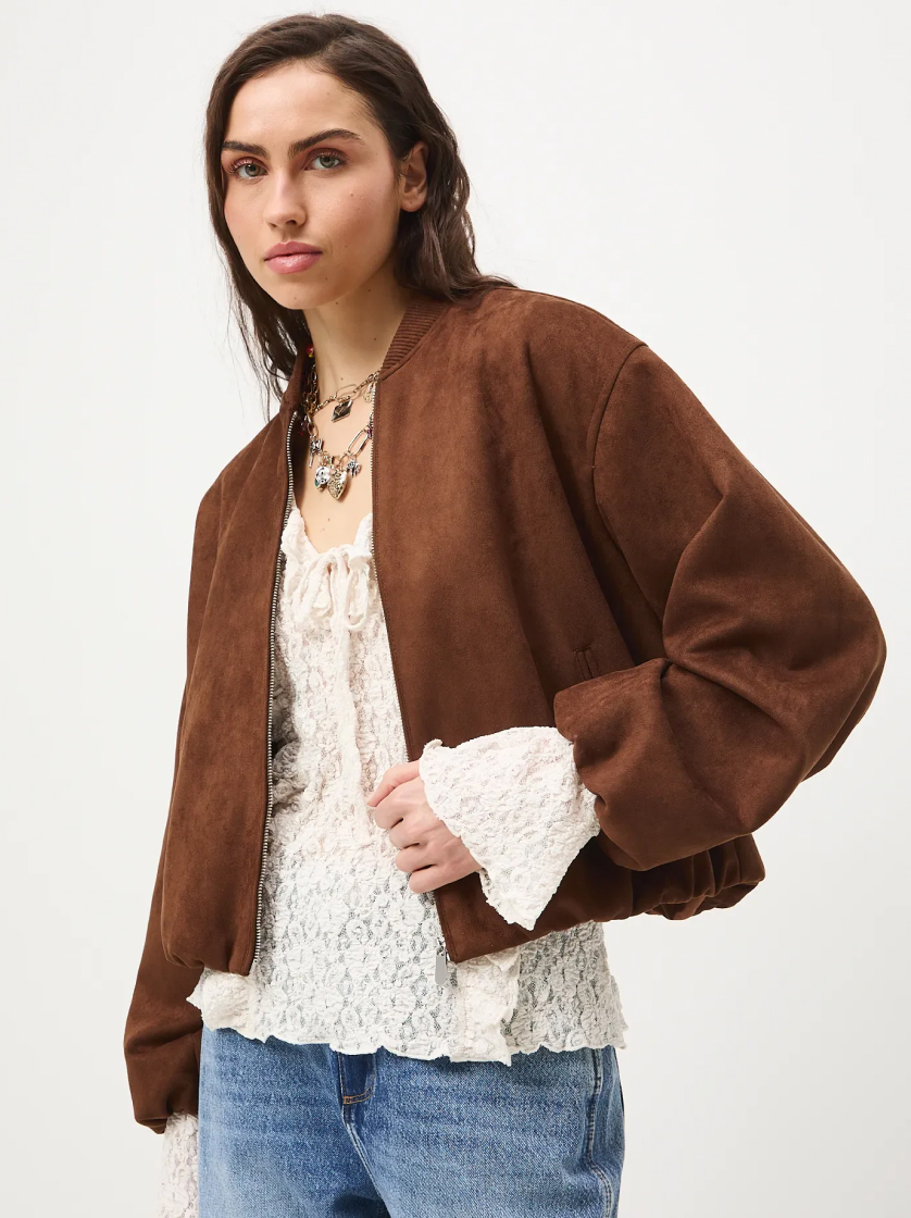 Modelka ubrana w krótką, brązową kurtkę zamszową typu bomber, jasną bluzkę z koronką i niebieskie jeansy, pozująca na białym tle.