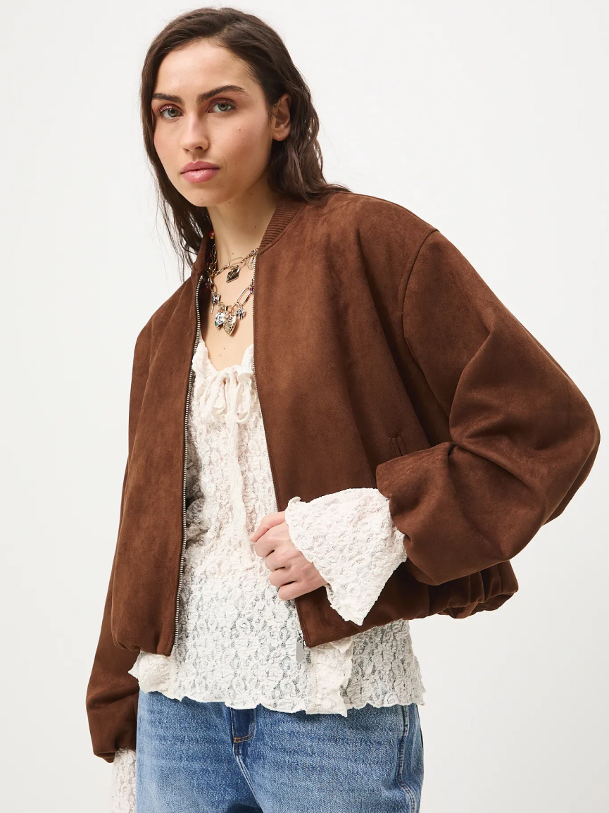 Modelka ubrana w krótką, brązową kurtkę zamszową typu bomber, jasną bluzkę z koronką i niebieskie jeansy, pozująca na białym tle.